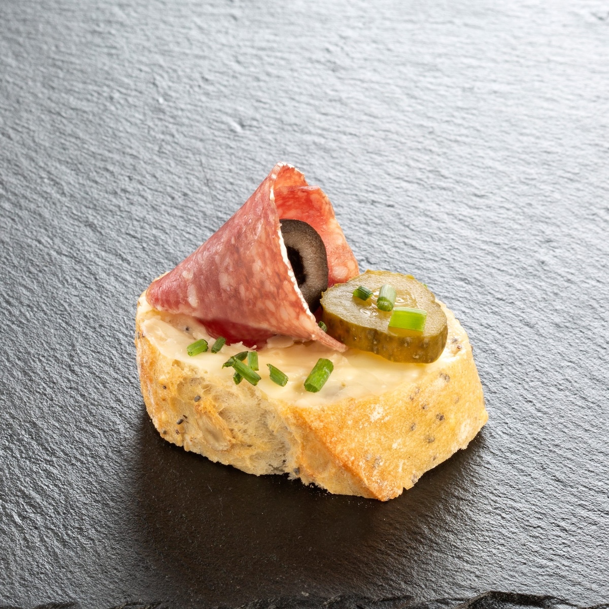 Salami-Häppchen auf Schiefer angerichtet: Toast mit Frischkäse, eingerollter Salami, schwarzer Olive und Gewürzgurke – Catering-Spezialität für Firmenevents in München